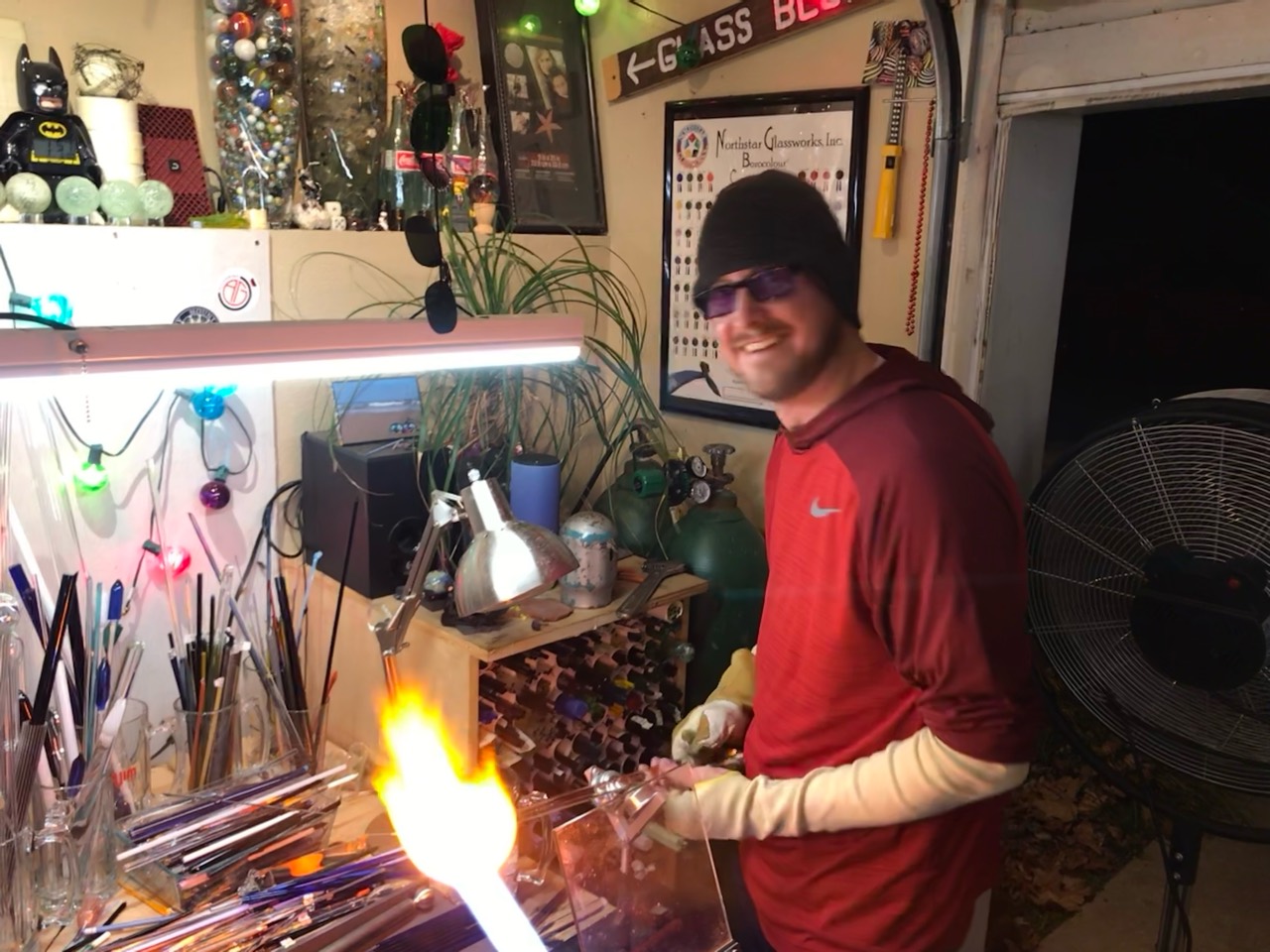 Will Stuckenberg smiling at his glassworking bench, surrounded by rods, tools and a lit torch