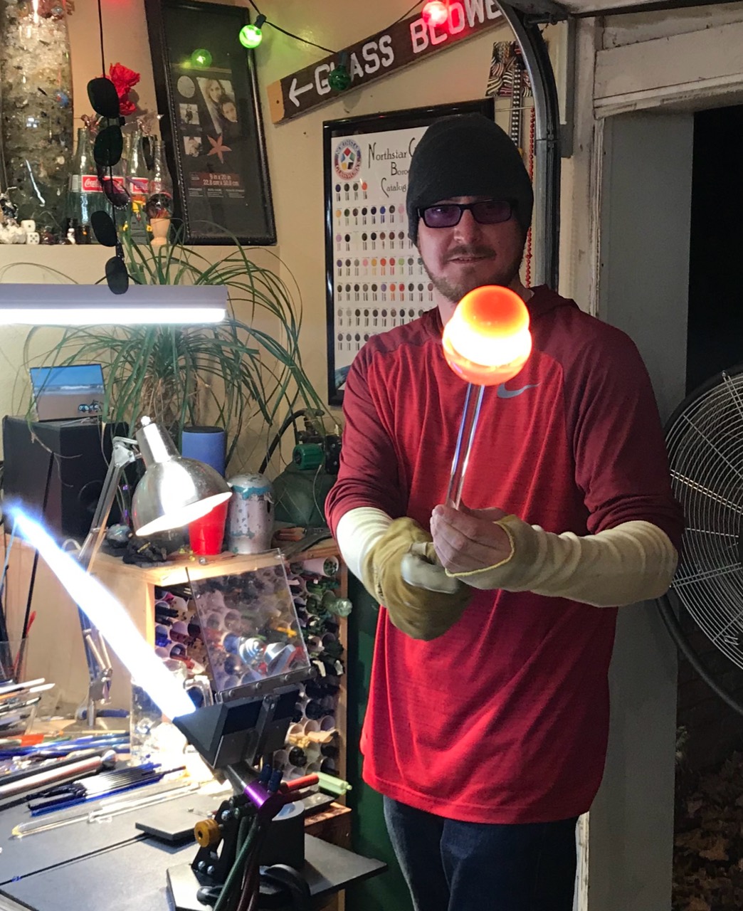 Will Stuckenberg holding a glowing glass piece on a rod in front of his workbench
