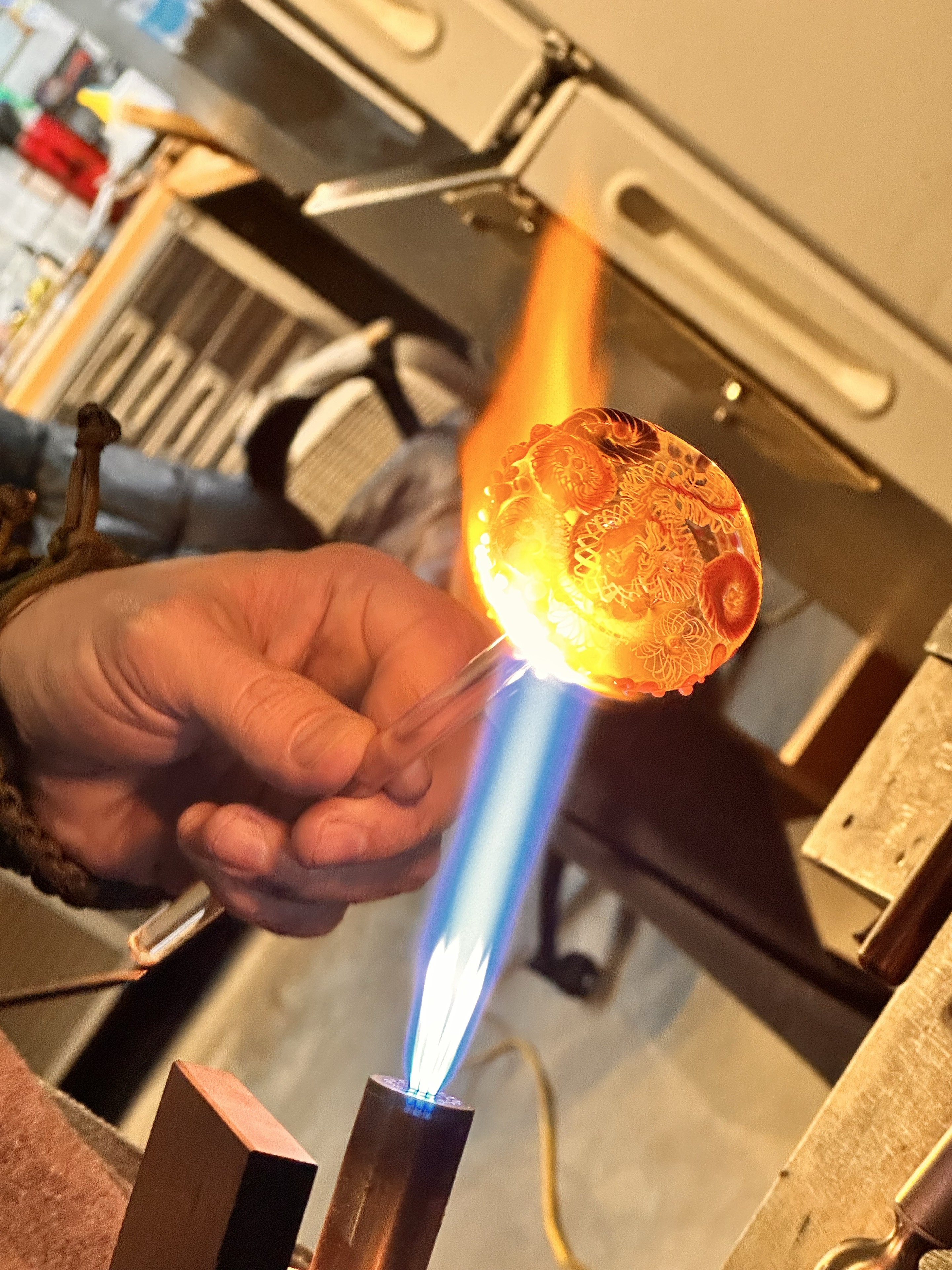 A hand holding a rod with intricate glowing glass over a blue torch flame