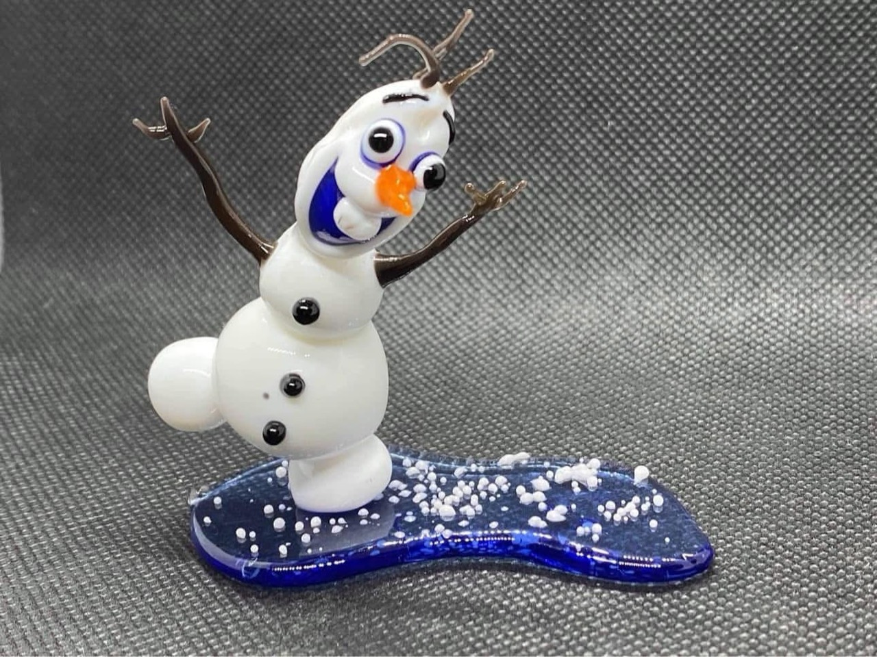 Olaf glass sculpture with raised twig arms, an orange nose and a snowy blue glass base