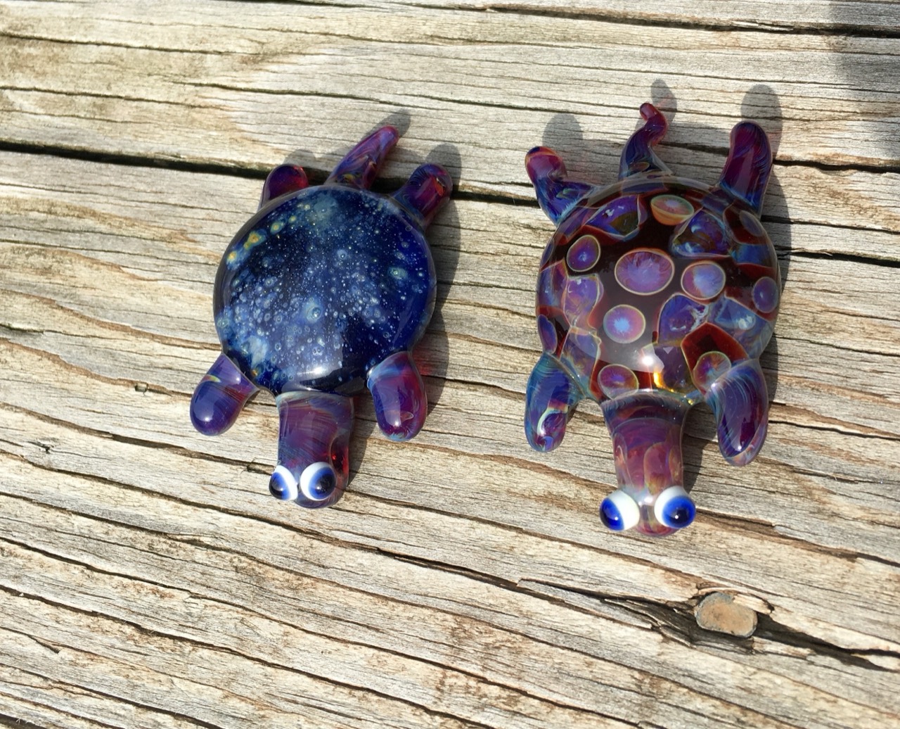 Cosmic Glass Turtles: Sunlit view of two glass turtles on weathered wood with wide blue-and-white eyes and colorful flippers