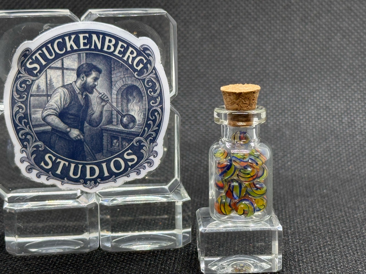 Electric Rainbow Mini Micro Marbles in their corked jar on a glass display stand beside studio branding in ordinary light