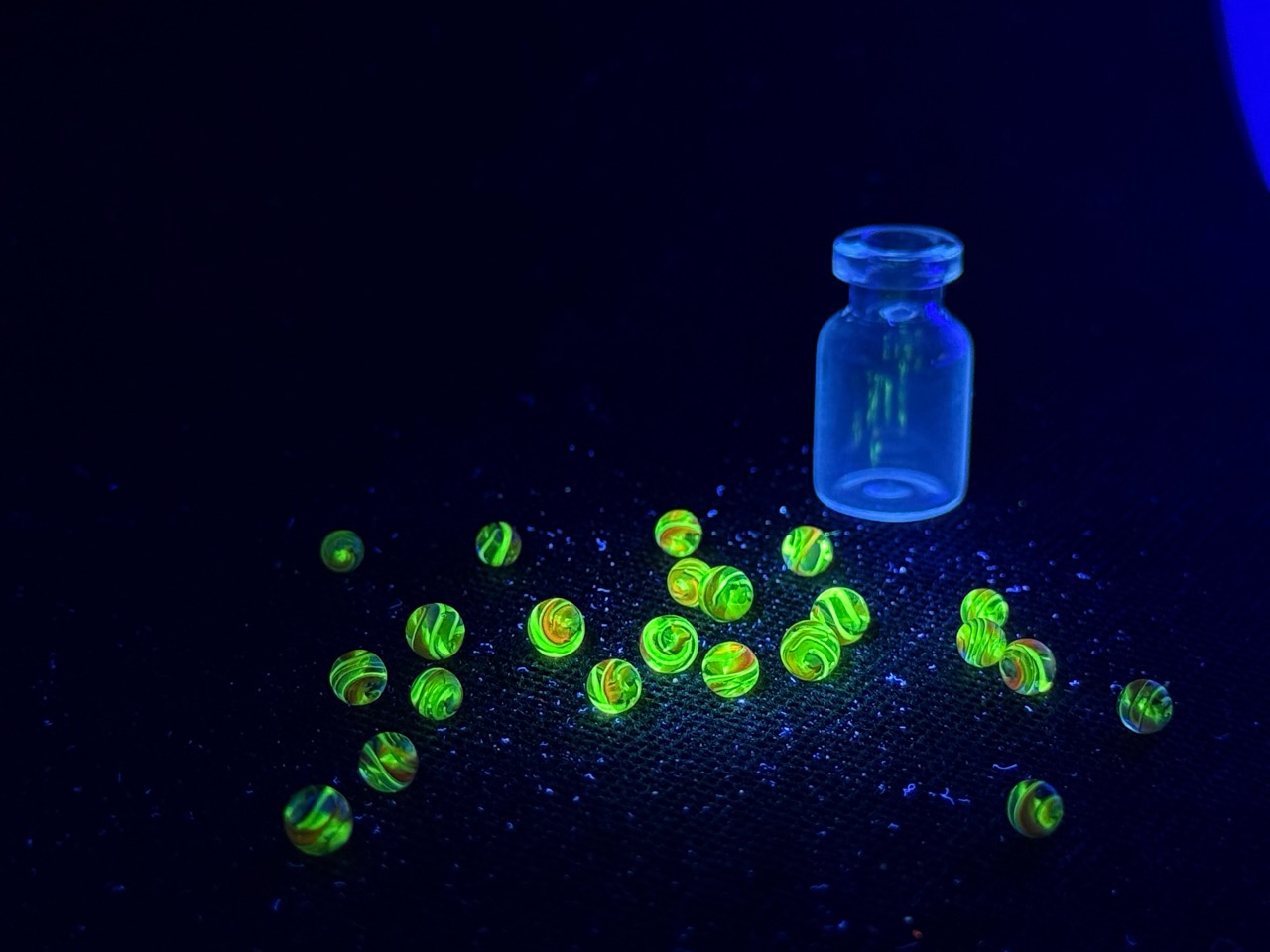 Wide view of Electric Rainbow Mini Micro Marbles glowing green under UV light beside an upright empty jar