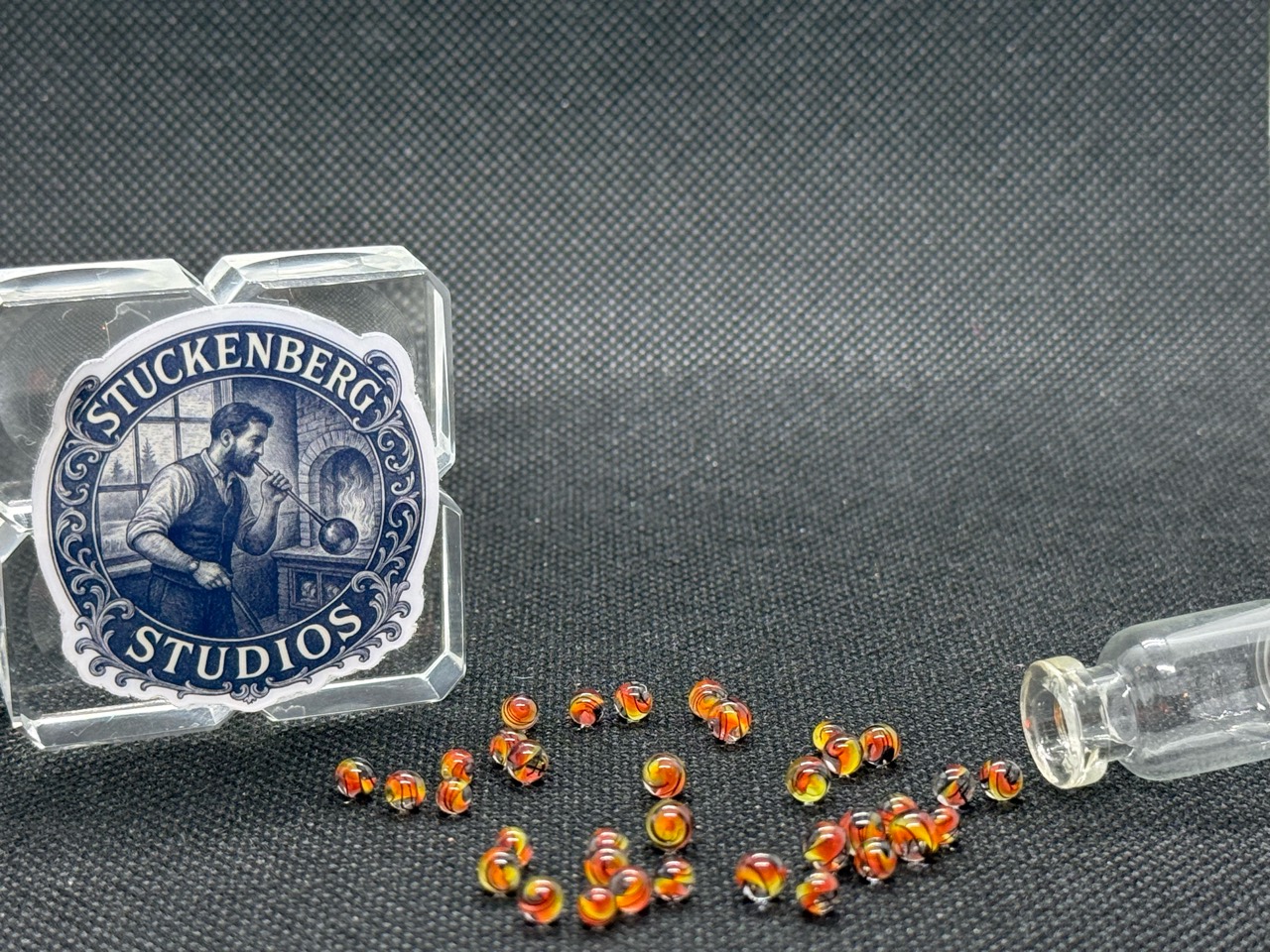Wide view of Fine Line Flame Mini Micro Marbles spread beside studio branding and their open jar