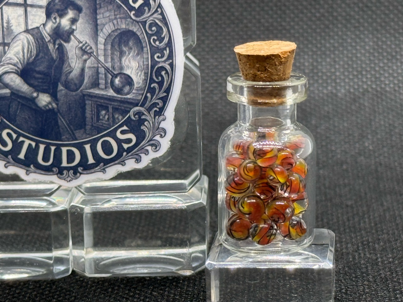 Alternate close view of Fine Line Flame Mini Micro Marbles in their corked jar beside studio branding