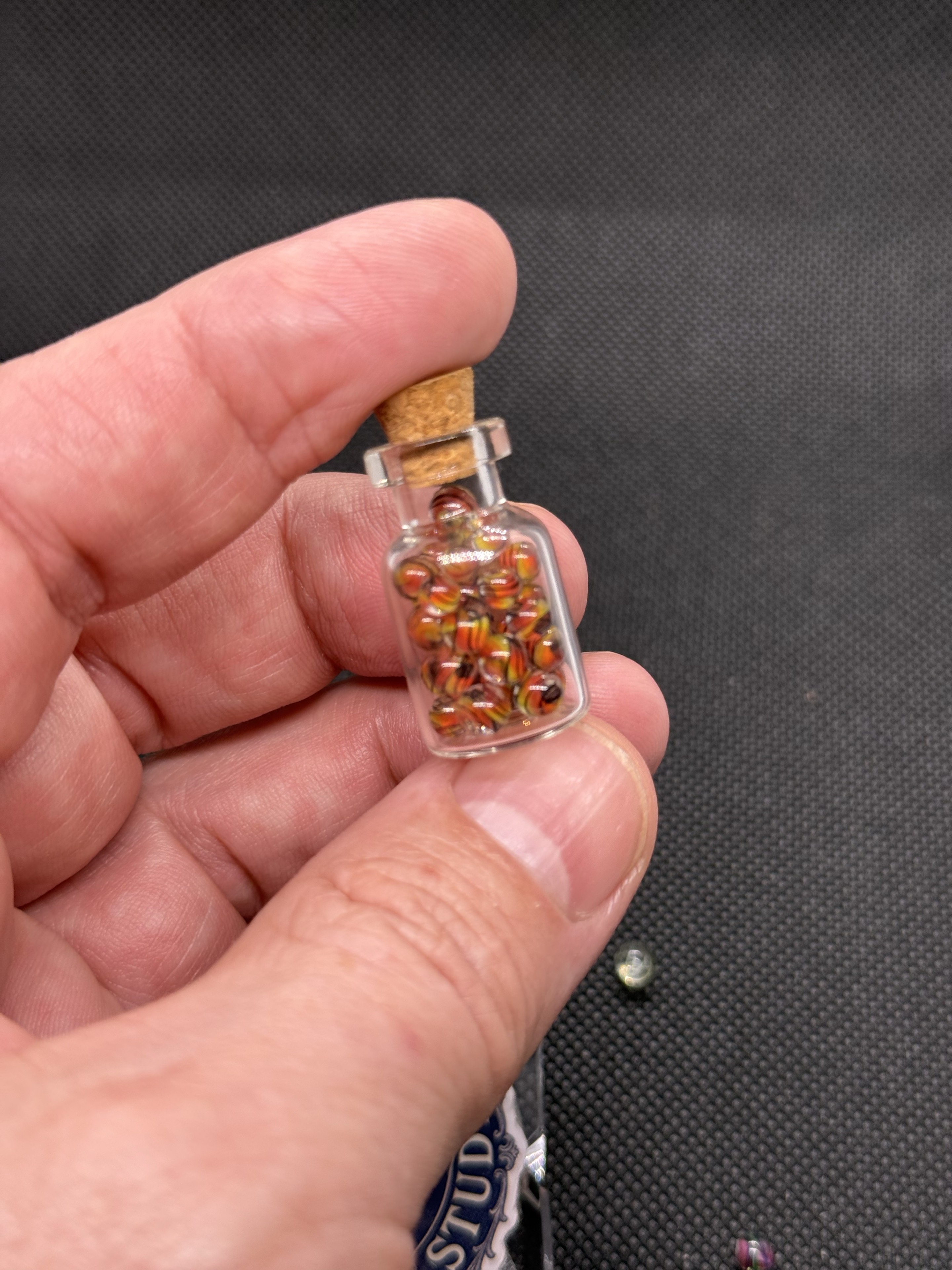 Fine Line Flame Mini Micro Marbles in a corked jar held between fingers for scale