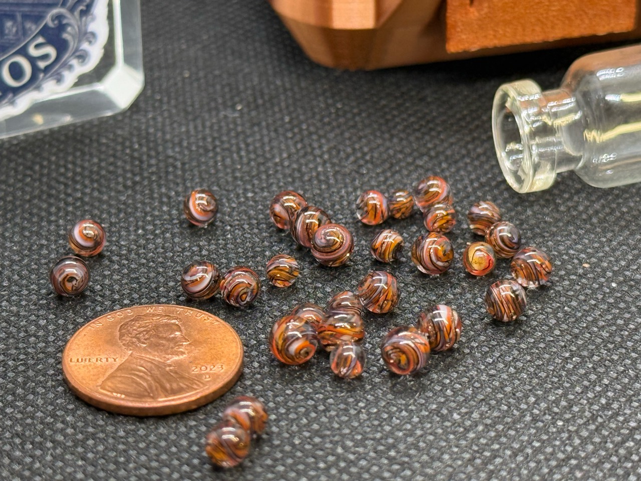 Wider view of Goldstone Mini Micro Marbles beside a penny and their open jar for scale