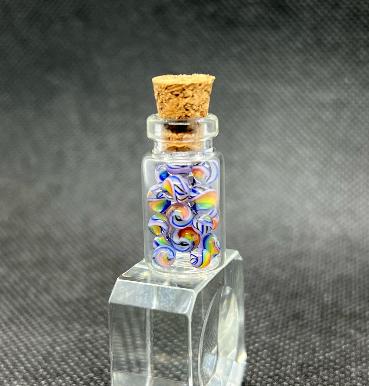 Legacy White Cloud Rainbow Mini Micro Marbles: Corked bottle of White Cloud Rainbow Mini Micro Marbles on a clear display block