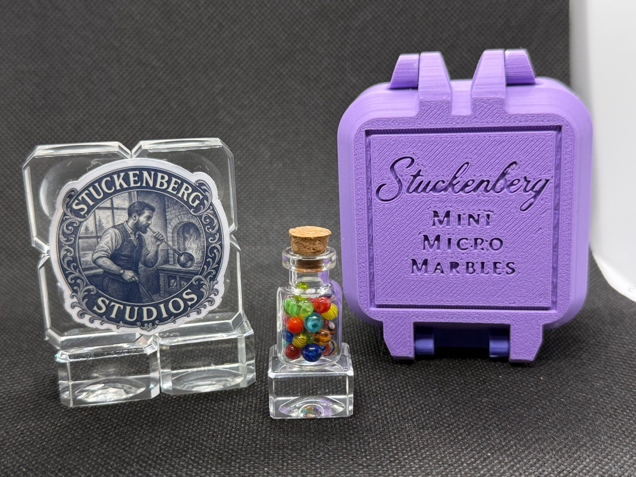 Mixed Colors Mini Micro Marbles in a corked jar beside studio branding and an upright lavender hard case