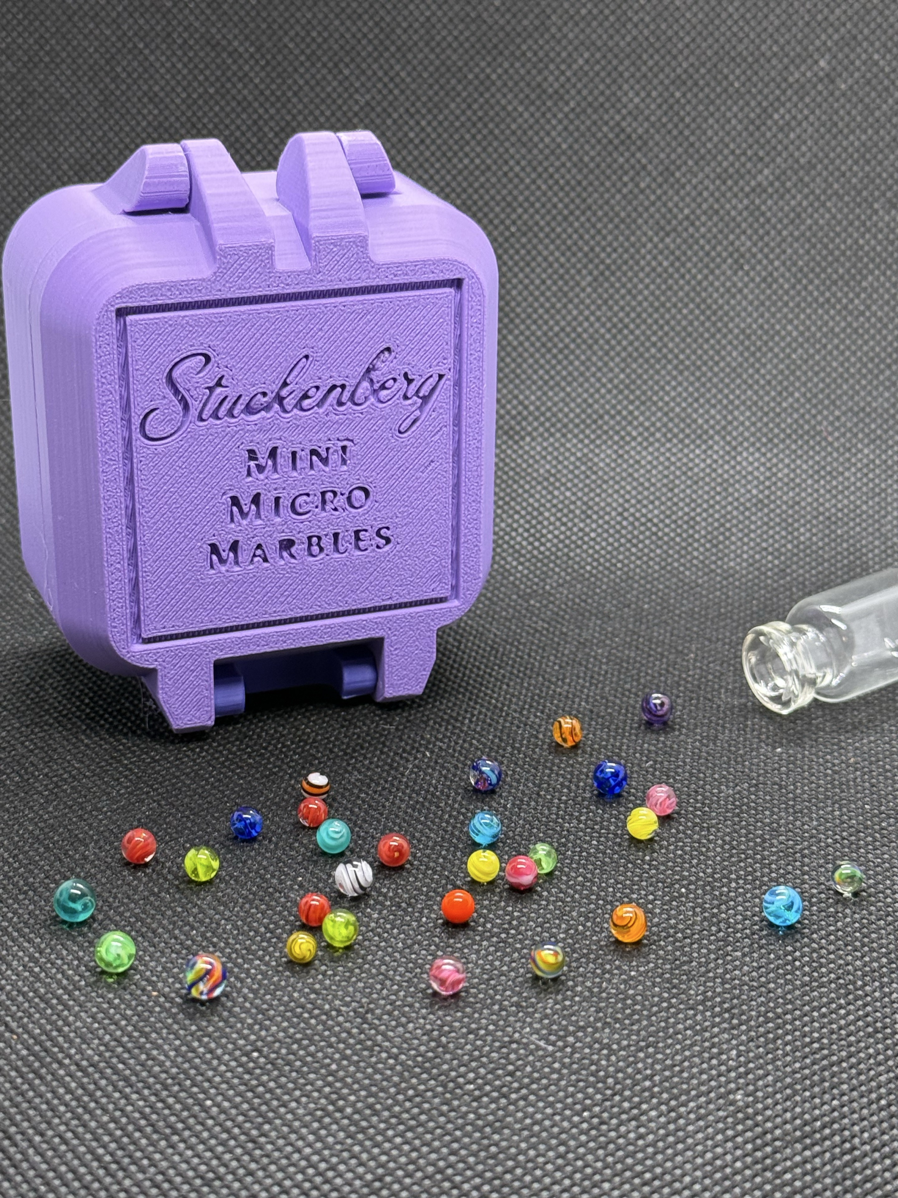 Portrait view of loose Mixed Colors Mini Micro Marbles beside their lavender hard case and open jar
