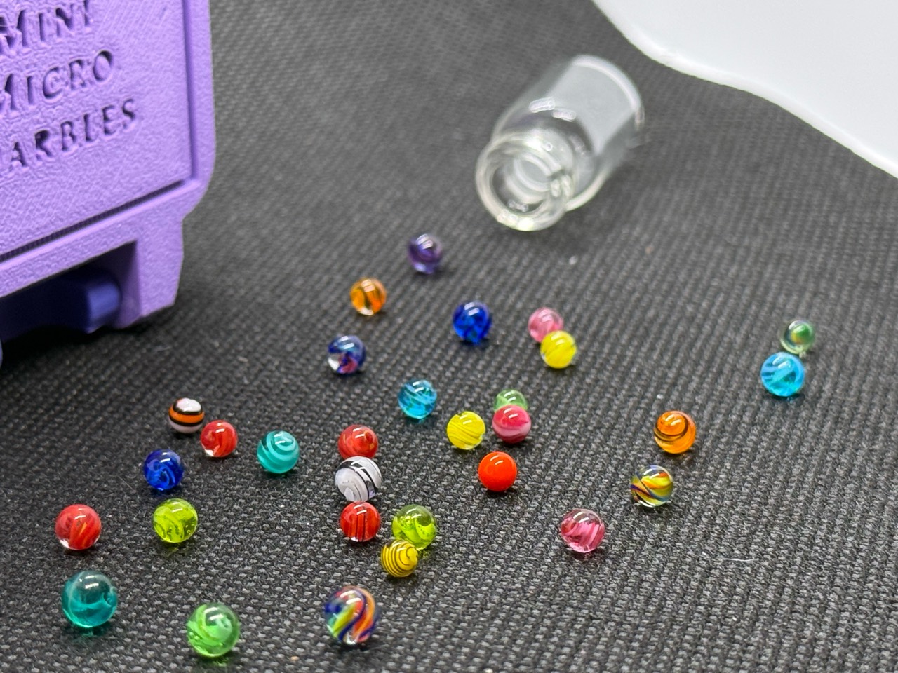 Mixed Colors Mini Micro Marbles spread in front of their open jar and lavender hard case