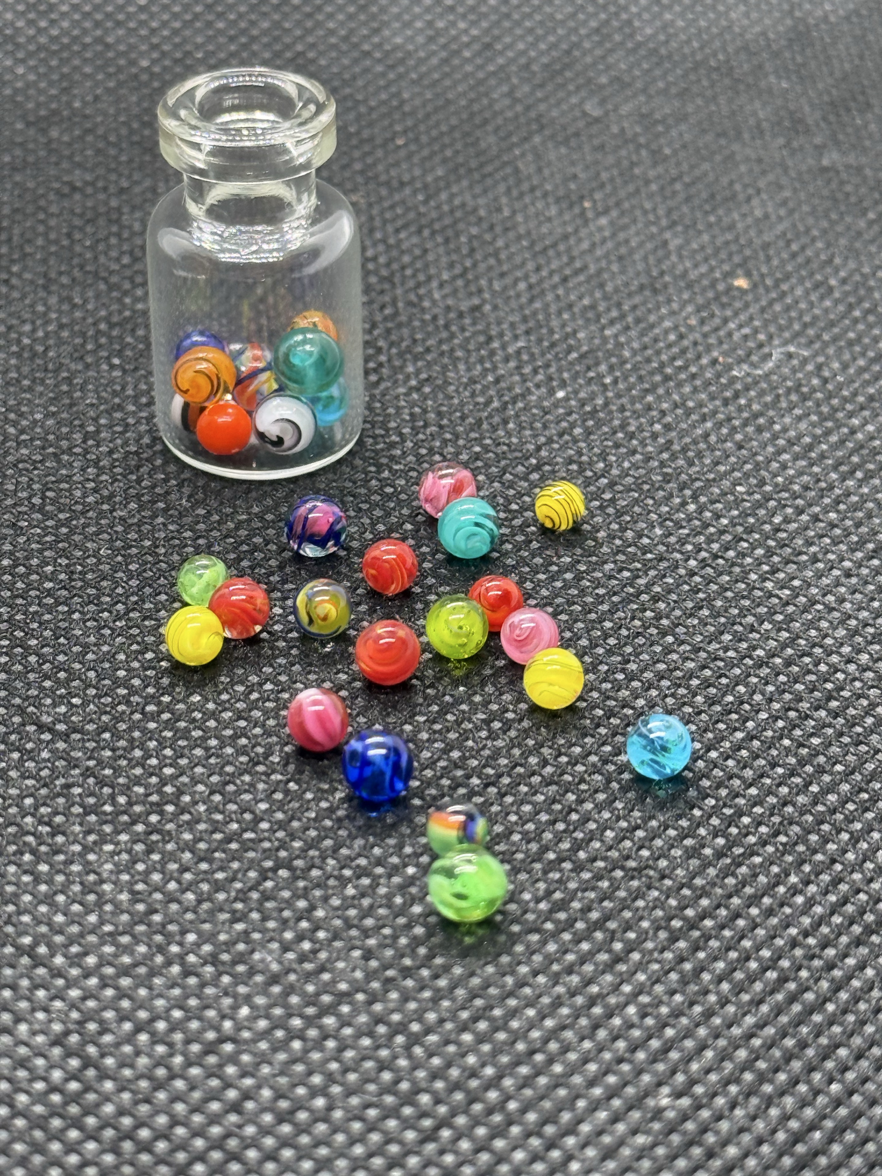 Mixed Colors Mini Micro Marbles spilling from an upright open jar onto dark fabric