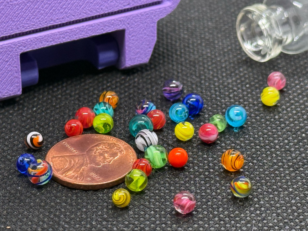 Close view of Mixed Colors Mini Micro Marbles around a penny for scale beside their open jar and lavender case