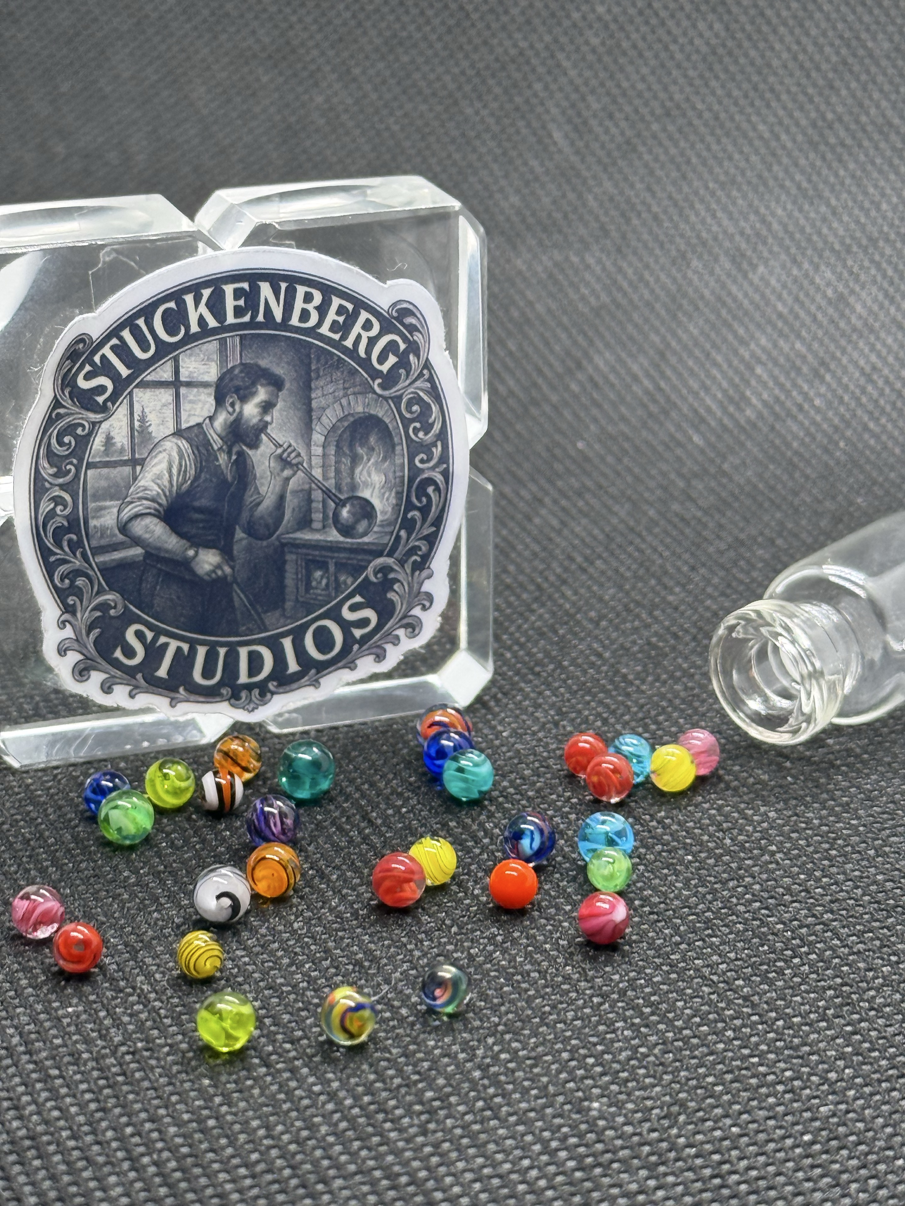 Portrait view of Mixed Colors Mini Micro Marbles grouped beside studio branding and their open jar