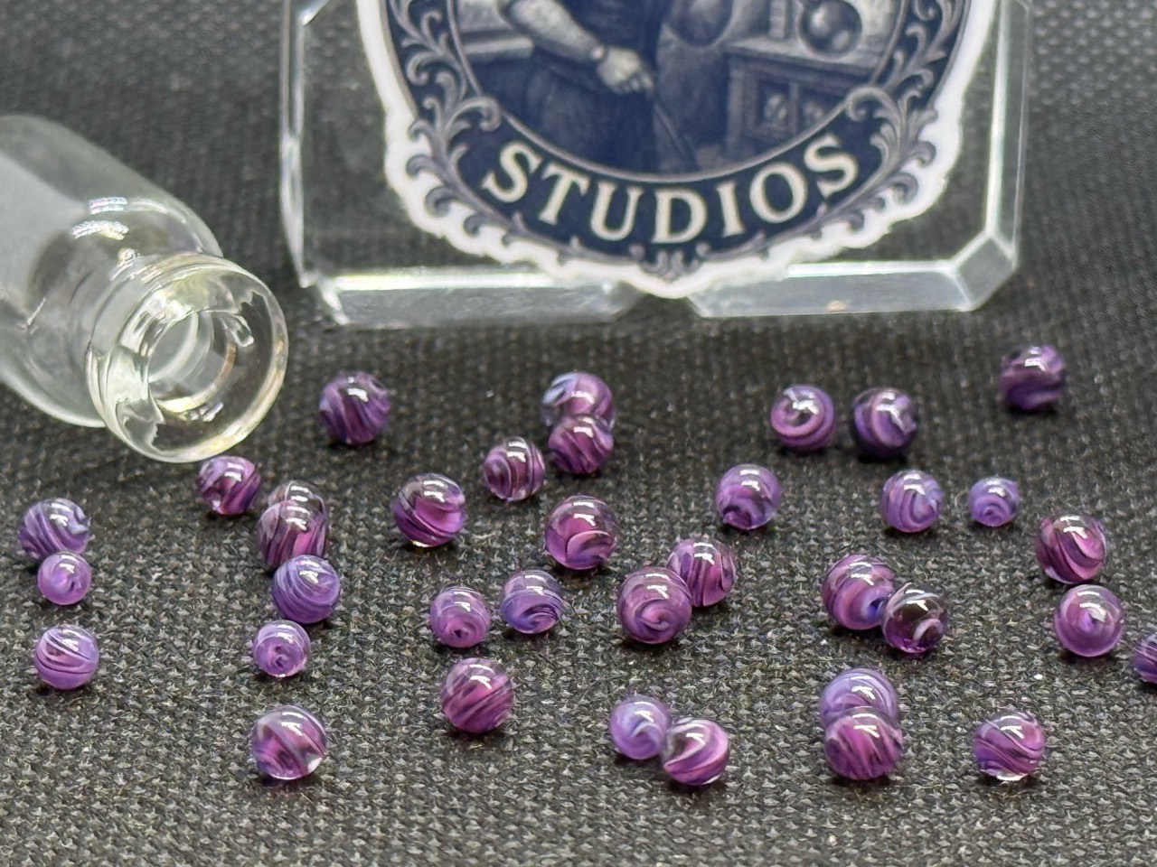Purple Dream Mini Micro Marbles with flowing purple ribbons and fine dark lines beside their jar