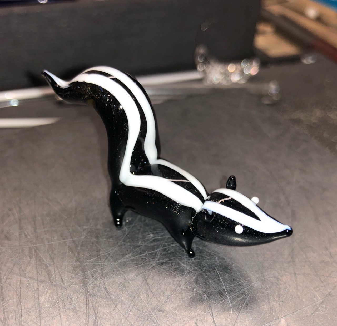 Sparkling Skunk Sculpture: Three-quarter view of the skunk on a metal work surface, showing paired white stripes and glittering black glass
