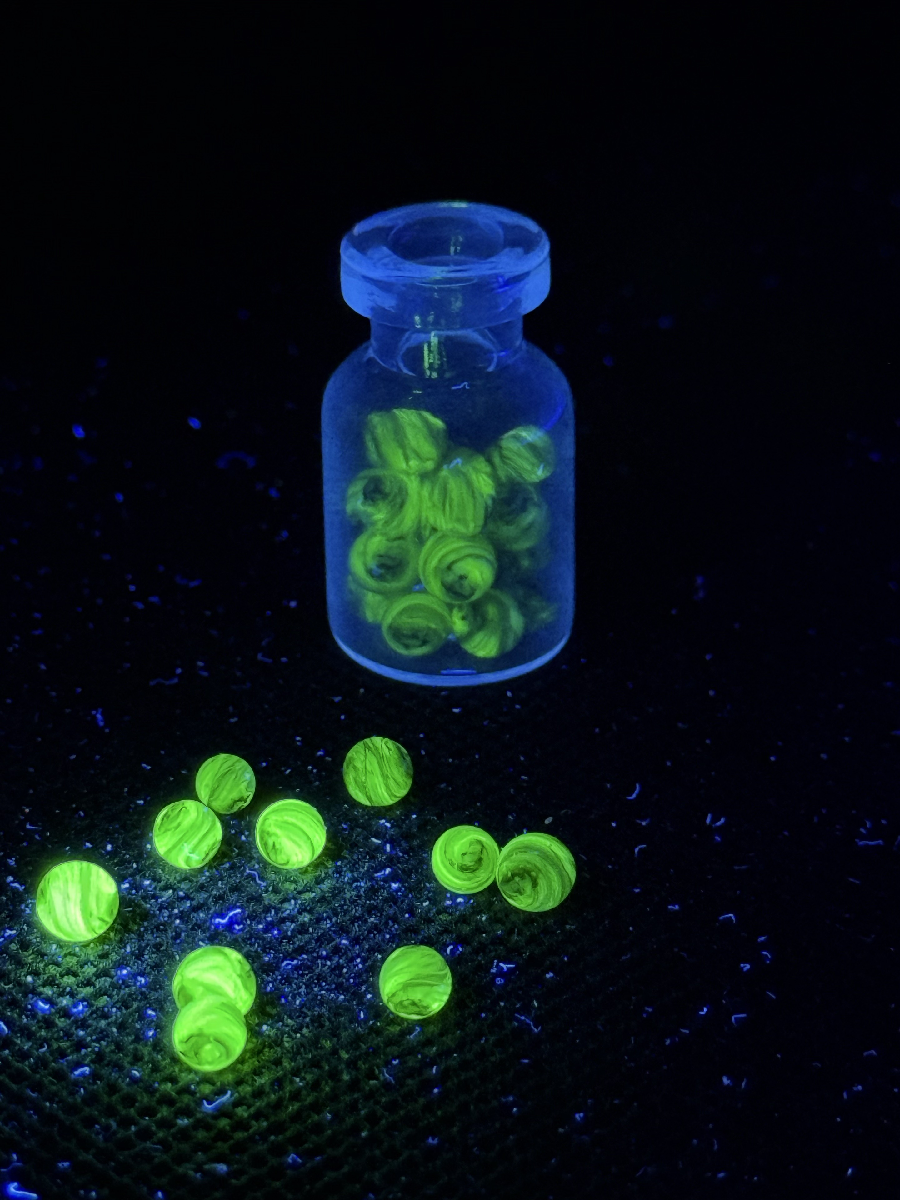 UV Champagne Mini Micro Marbles glowing green in and around their open jar under UV light