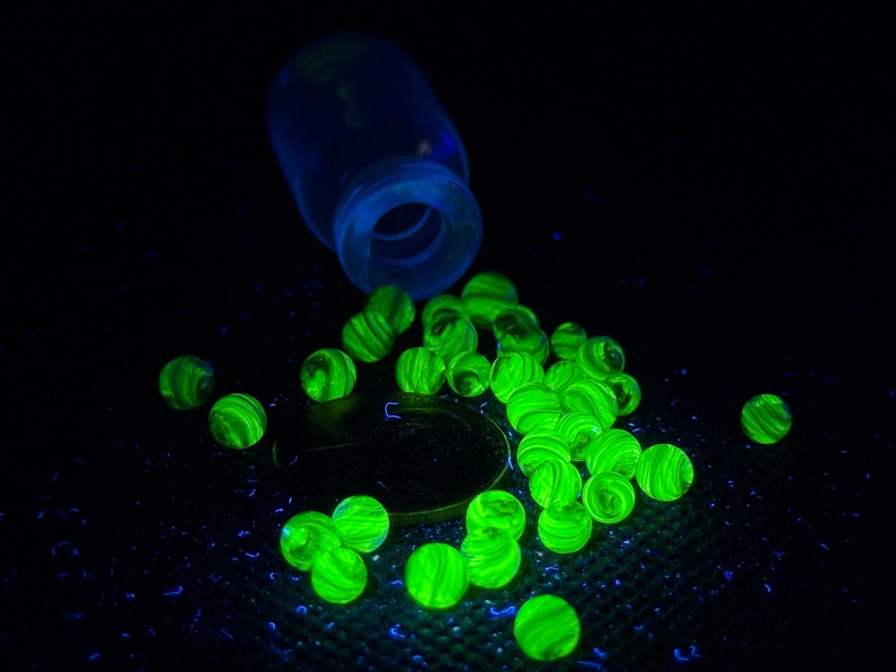 UV Champagne Mini Micro Marbles glowing green around a penny beside their open jar under UV light for scale