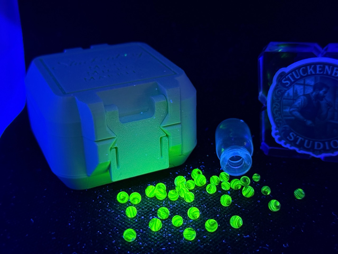 Wide view of UV Champagne Mini Micro Marbles glowing green beside their open jar, pale case and studio branding under UV light