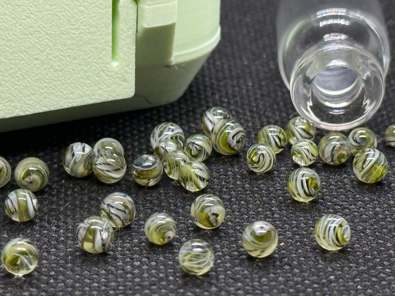 Close view of fine lines and swirls in loose UV Champagne Mini Micro Marbles beside their open jar and hard case in ordinary light