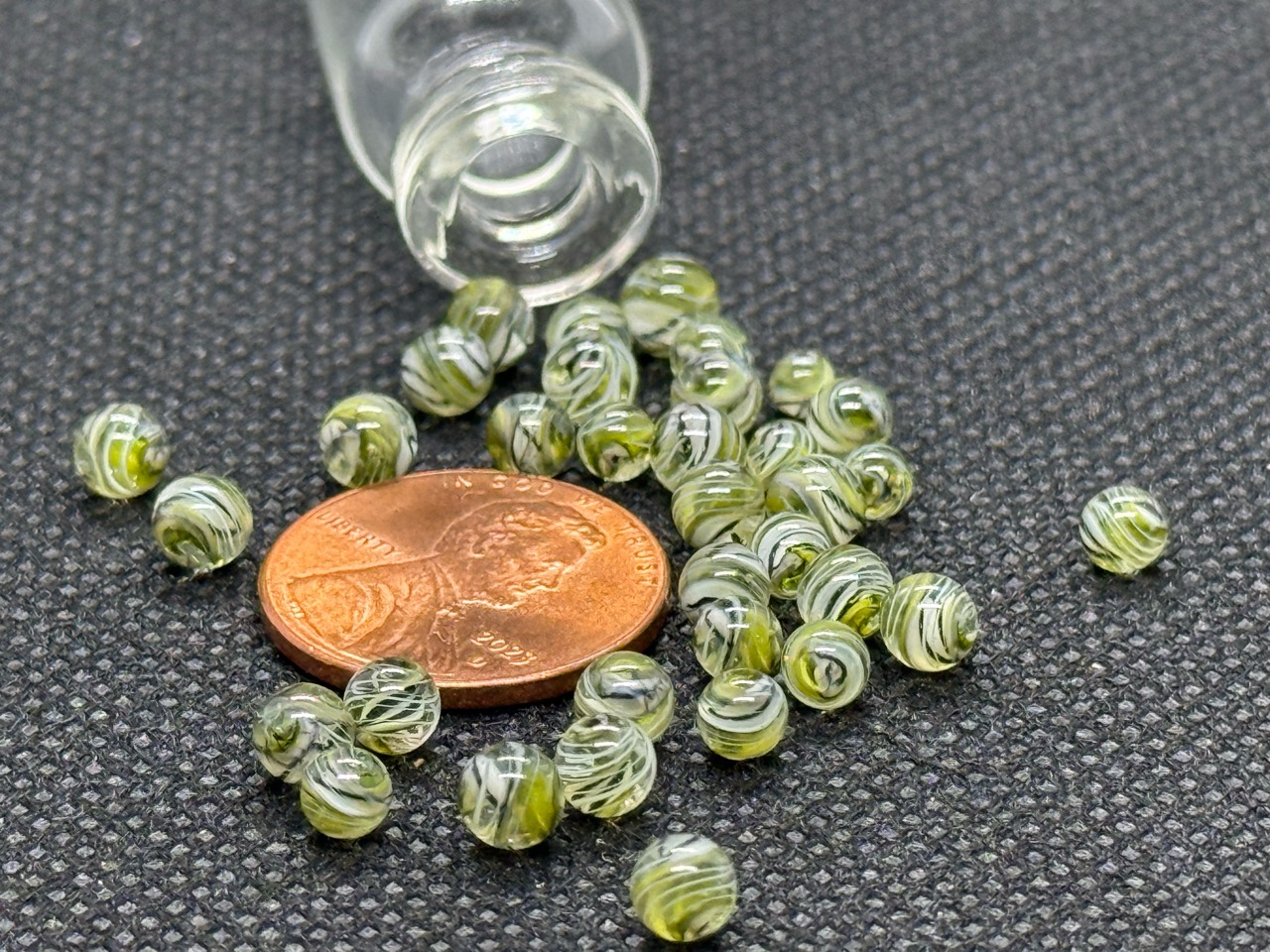 UV Champagne Mini Micro Marbles with fine lines spread around a penny beside their open jar in ordinary light for scale