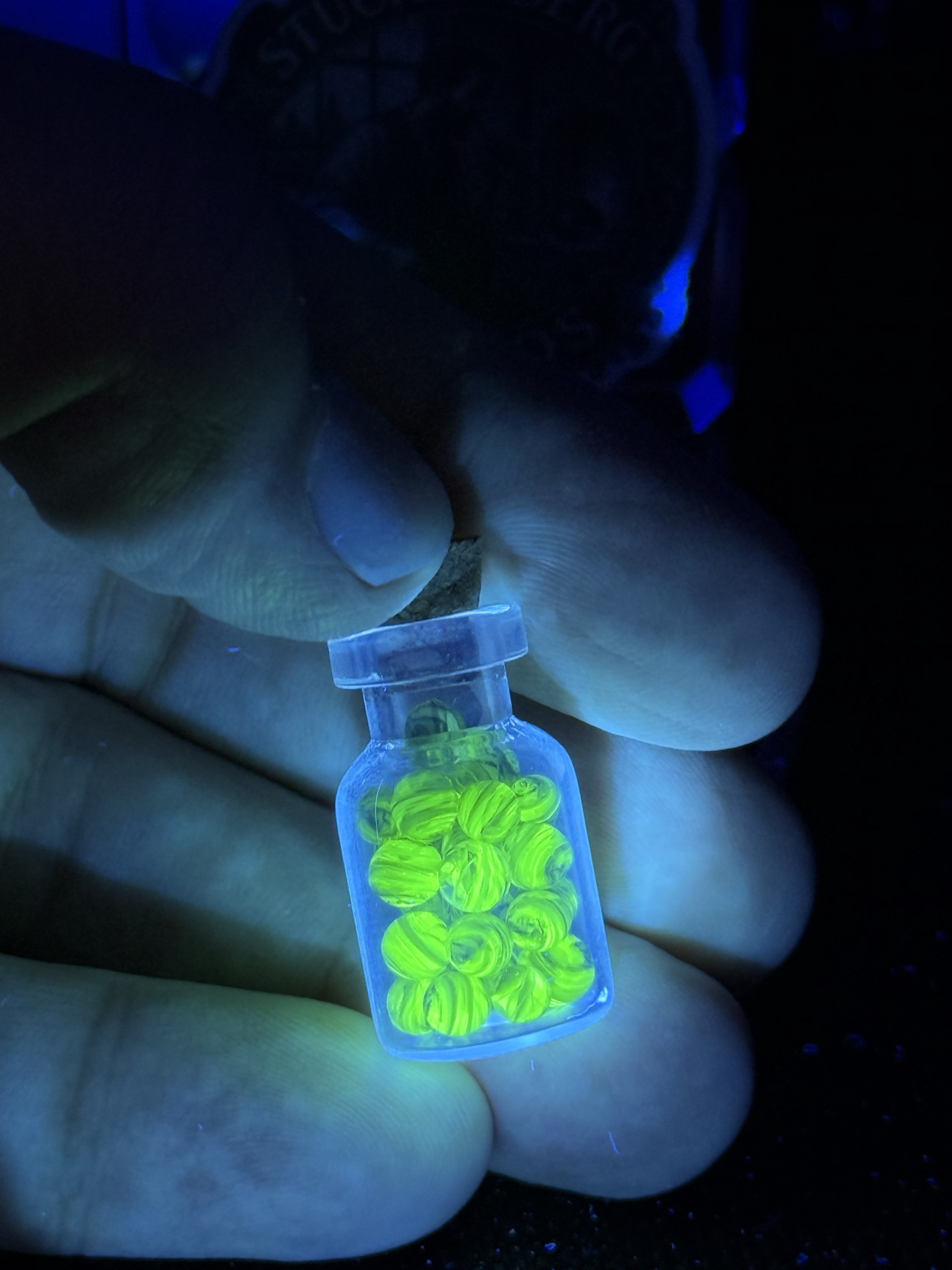 Jar of UV Champagne Mini Micro Marbles glowing bright green under UV light while held in hand for scale